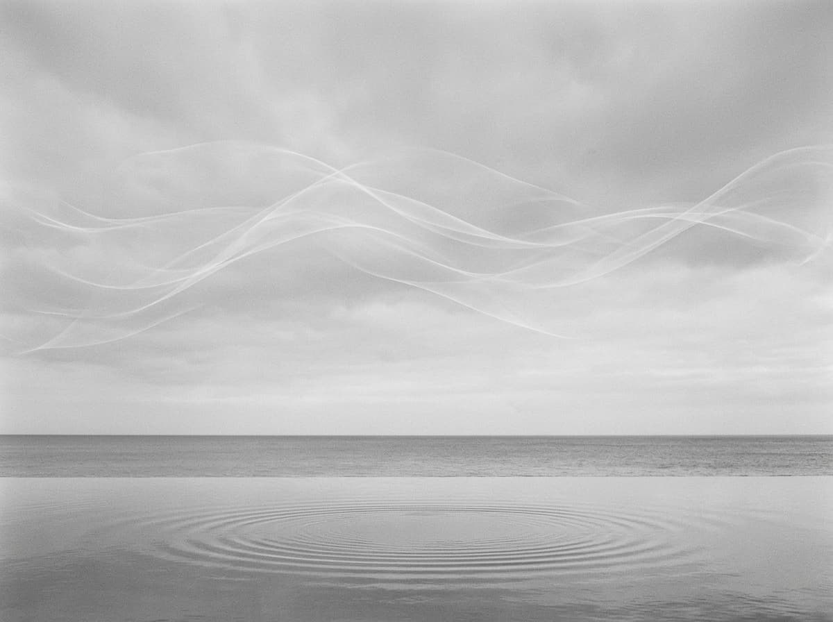Monochrome seascape: translucent ribbons of wind above the horizon and a ripple spreading across still water